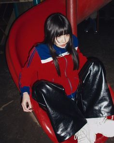 a woman sitting on top of a red chair next to a metal pole and wearing black leather pants