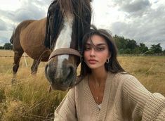 a woman standing next to a horse in a field with tall grass and cloudy skies