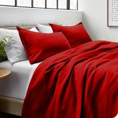 a bed with red sheets and pillows in a white room next to a plant on a table