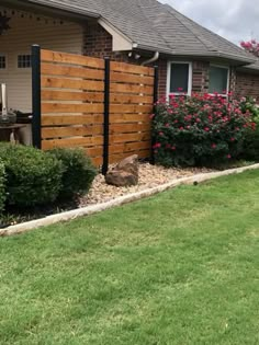 a wooden fence in front of a house with flowers on the lawn and bushes around it