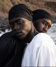 two black men standing next to each other near the water and rocks in the background