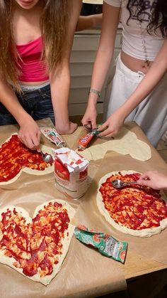 two girls are making heart shaped pizzas on a table with cans of pepperoni