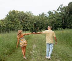 a man and woman holding hands walking down a path in the grass with trees behind them