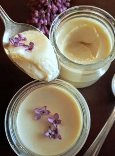 two jars filled with cream and flowers on top of a table next to spoons