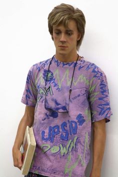 a young man standing against a white wall holding a book and wearing a purple t - shirt