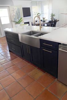 a kitchen with black cabinets and white counter tops, an island in the middle is surrounded by brown tile flooring