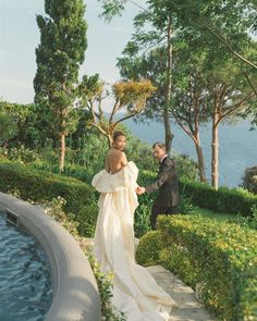 a man and woman standing next to each other in front of a garden with water