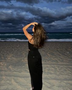 a woman standing on top of a sandy beach next to the ocean under a cloudy sky