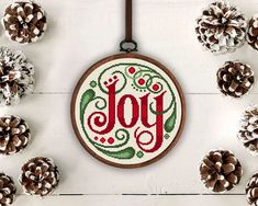 a cross stitch christmas ornament surrounded by pine cones and fircons with the word joy written in it