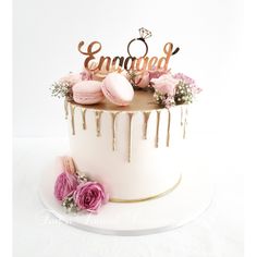 a white cake with pink flowers on top and the word engaged spelled in gold letters