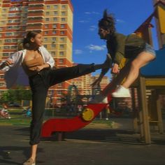 two people doing tricks on a skateboard at a park in front of tall buildings