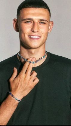 a young man wearing a necklace and bracelet with his hands on his chest smiling at the camera