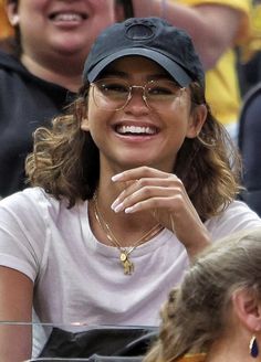 a woman wearing glasses and a baseball cap sitting in the stands at a tennis match