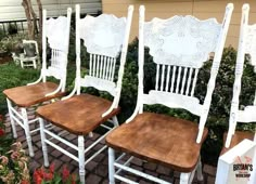 three white chairs with wooden seats sitting in front of some bushes and flowers on the ground