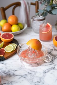 grapefruit punch being poured into a glass pitcher on a marble table with citrus fruit in the background