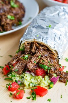 shredded beef wrapped in tin foil with vegetables and tomatoes on the side next to a bowl of salad