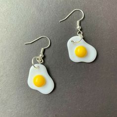 two fried eggs are hanging from silver earwires on a gray surface, one has a yellow egg in the middle