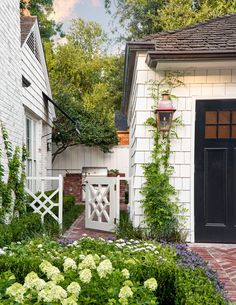 a white house with a black door surrounded by greenery and flowers in front of it