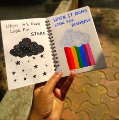 someone holding up a notebook with an image of a rainbow and clouds on it that says when it's dark look for stars