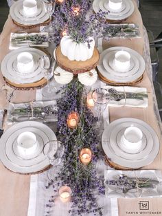 the table is set with white plates, silverware and lavender flowers in vases