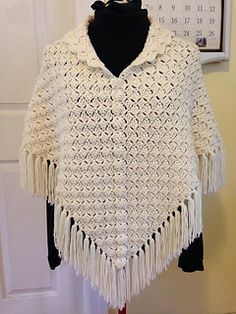 a white crocheted jacket with fringes hanging on a wall next to a black mannequin