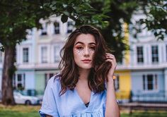 a woman with makeup on her face is standing in front of a tree and building