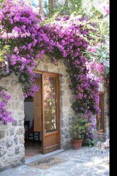 purple flowers are growing on the side of a stone building with an open door and glass windows