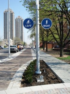 two blue street signs sitting on the side of a road next to trees and buildings