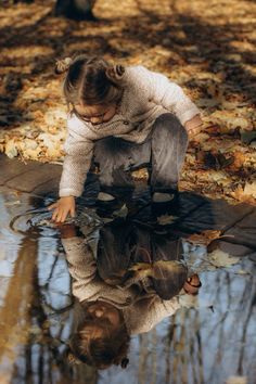 a woman kneeling down next to a puddle with leaves on the ground and trees in the background