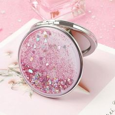 a compact mirror with glitter on it sitting on a pink surface next to a flower