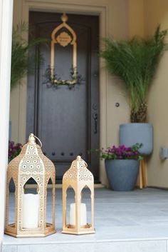 two white candles are sitting on the front porch with potted plants in the background