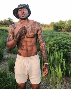 a shirtless man with tattoos on his chest and hands in his pockets, standing next to tall grass