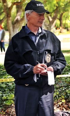 an older man wearing a police uniform standing in front of a park with trees and bushes