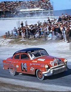 an old red car driving down a race track in front of a large group of people