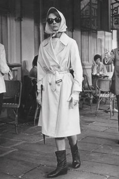 a woman in a trench coat and sunglasses is standing on the sidewalk with other people