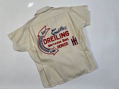 "Vintage 1950's Hilton bowling shirt w/ original tag. Never worn. Off white color. On the back: \" Bill Lark, Carillacs Dreiling, Mercedes Benz, Denver\" in blue and red flocked lettering. One chest pocket. Cuffed sleeves. There are a couple small stains on the front and a little bit of blue stain from the lettering -from being stored folded on itself. This shirt is still in great condition. Please see all photos and measurements, item is being sold as-is. . M E A S U R E M E N T S.  measured laying flat  Shoulder to shoulder: 15.5 inches  Pit to pit: 20.5 inches   Waist: 17.75 inches   Length: 23.5 inches  Sleeve length: 6 inches   Material:  rayon Size:  labeled 36 , fits like s/m Label:   Hilton Bowling shirts, Chicago  Please see Shop Policies before committing to purchase.   ALL SALES Vintage Racing Tshirt, Vintage Mechanic Shirt, Stained Clothes, Vintage Mechanics, Vintage Bowling Shirts, Vintage Shirt Design, Bowling Shirt, Blue Stain, Mechanic Shirts