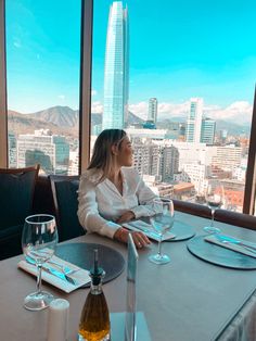 a woman sitting at a table with wine glasses in front of her looking out the window