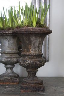 two metal vases with grass growing out of them on a table next to a mirror