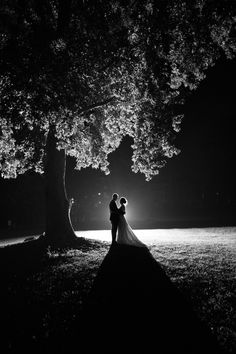 a bride and groom standing under a tree at night in black and white with their arms around each other