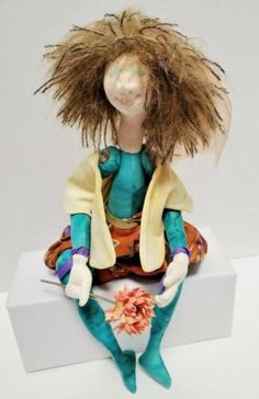 a doll with long hair sitting on top of a white box and holding a flower