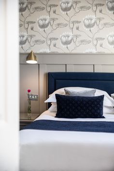 a bed with white sheets and blue headboard next to a flowered wallpaper