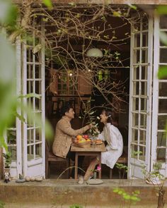 two people sitting at a table with fruit in front of an open door to a house