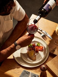 a person pouring wine into a glass on top of a plate with food and utensils