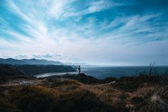 a person standing on top of a hill next to the ocean under a cloudy blue sky