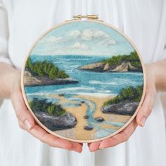 a person holding a small embroidery project with water and land in the background on a white shirt