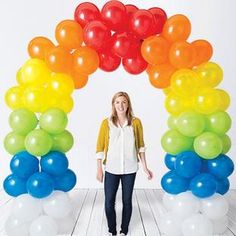 a woman standing in front of a rainbow arch with balloons on the sides and an orange, yellow, green, blue, red, and white balloon