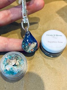 a hand holding a small glass container with sea shells in it next to a keychain