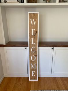 a wooden welcome sign sitting on top of a hard wood floor next to white cabinets