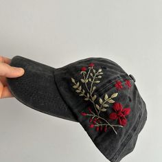 a hand holding a black hat with red flowers and leaves on the front, while it is held up against a white wall