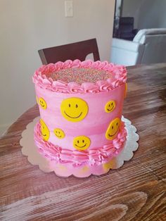 a pink cake with smiley faces on it sitting on top of a wooden table next to a chair